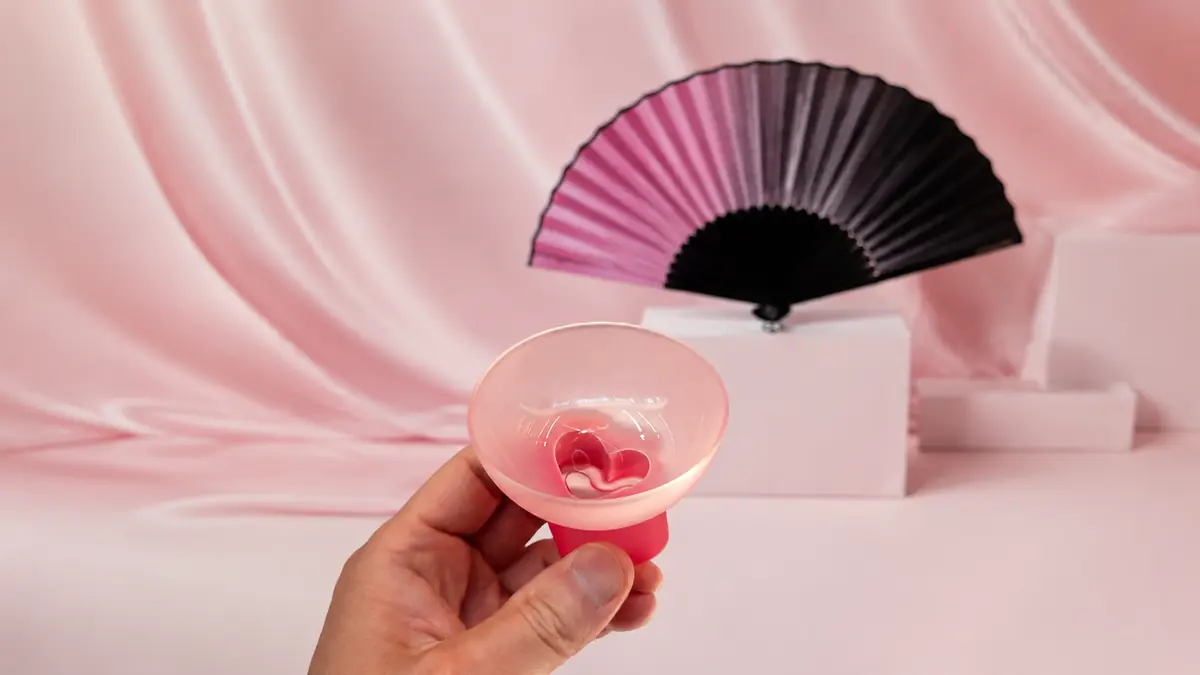 A hand holding a frosted pink BLACKPINK × MUD:S soju glass with a pink-to-black folding fan in the background